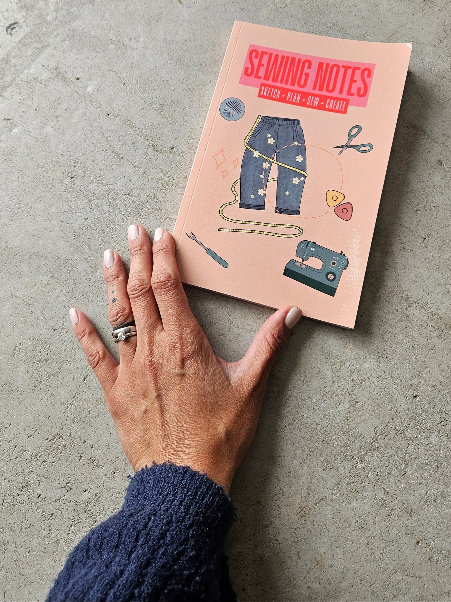 Hand holding a pink sewing themed notebook with illustrations on a concrete floored